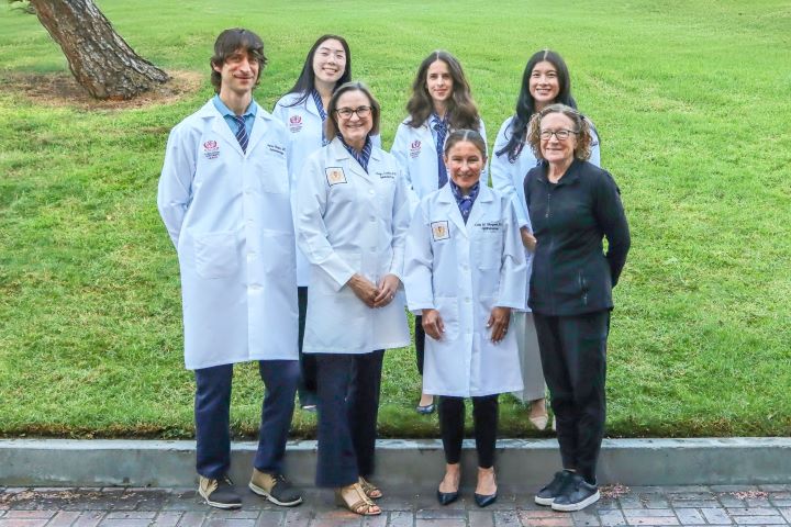 Photograph of six people standing outdoors on a paved area with grass in the background, five wearing white lab coats and one dressed in black casual attire. The group appears to be medical or scientific professionals, indicated by lab coats and identification badges, suggesting a team or staff photo.