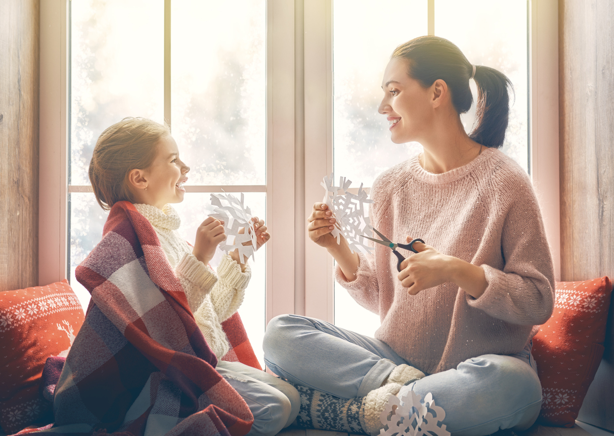 female child and female adult cut snowflakes in front of snowy window