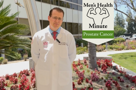 A doctor wearing a white coat stands with his hands behind his back while looking into the camera. He is in front of a medical building