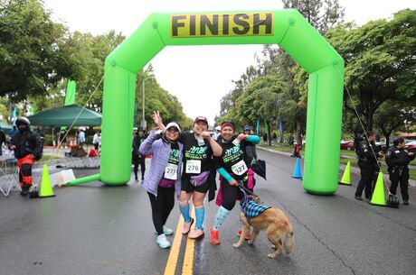 Three female participants and a dog stand in front of the green finish line holding their race medals