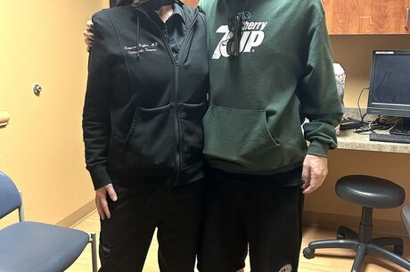 A female doctor in all black stands next to her male patient, who wears a green sweatshirt and black shorts. They are in a doctors office that has yellow walls