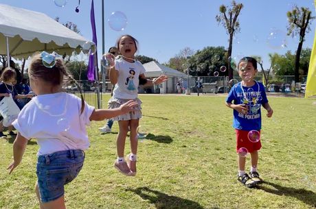 Three children in tee-shirts and shorts play outside in the grass blowing bubbles and smiling 