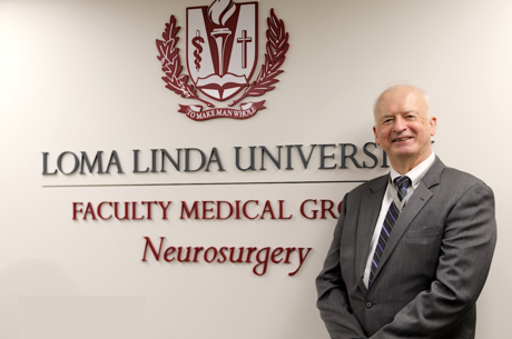 Man stands in front of a white wall with a sign saying Loma Linda University Faculty Medical Group Neurosurgery