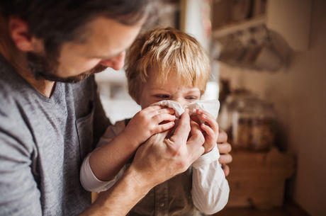 A father on paternity leave looking after small son indoors, blowing his nose