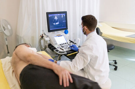 Doctor looking at the computer screen while doing an ultrasound test of a senior male patient lying on clinic bed during pandemic. Cardiologist doing heart ultrasound of an elderly man in hospital.