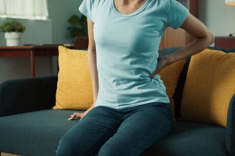 Persistent aches and pains could indicate something more serious Woman hand holding her low back, feeling low back pain and hurt while sits on sofa in living room at home