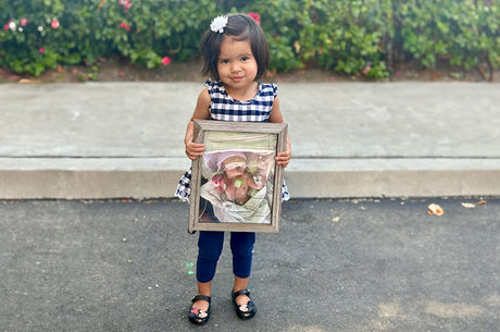 Little girl wearing black and white dress with leggings and flats holds a photo of herself when she was in the NICU