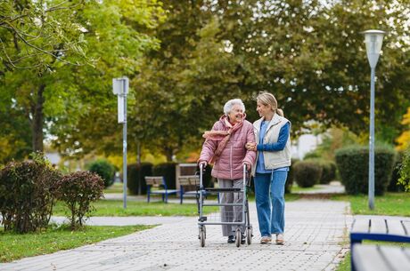 Senior woman and her home caregiver spending a chilly, windy day outdoors in city park. Autumn walk for elderly patient with walker