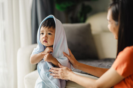 Asian boy with a towel on his head is sitting in the living room. He has big, dark black eyes and is relaxing on the sofa after his bath.