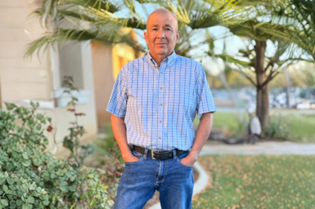Man in blue button up and jeans stands in front of his house with his hands in his pocket