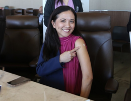 A women smiles at the camera and points to a bandaid on her arm 
