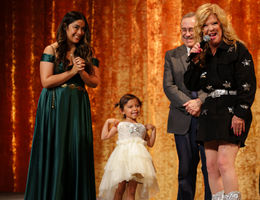 two women and a man on stage at a gala, with a young girl wearing an ivory dress a cowboy boots