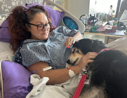 Woman lays in a hospital bed with glasses on and a black and dog medium sized dog laying across her lap.