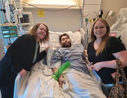 Two woman lean over a man in a hospital bed while all three smile at the camera