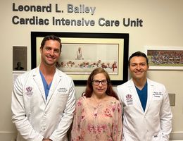 Colleen Barber reunites with the structural interventional cardiologists who recently performed a procedure to close a hole in her heart, Dr. Jason Hoff (left) and Dr. Amr Mohsen (right), framed by a background of photographs of Dr. Leonard Bailey who transplanted Barber&#039;s heart when she was a baby.