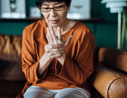 Senior Asian woman rubbing her hands in discomfort, suffering from arthritis in her hand while sitting on sofa at home